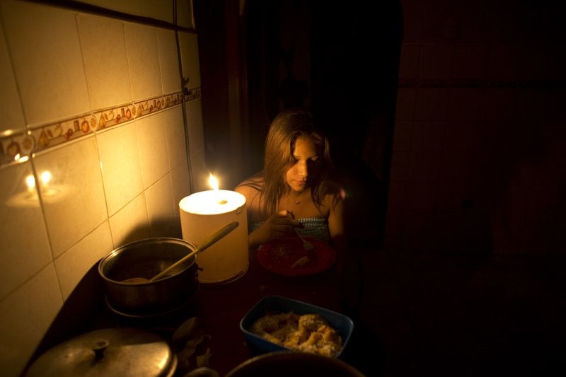 A woman eats in a dark kitchen next to a lit candle. 