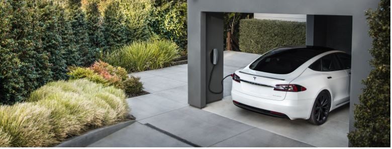 A Tesla car sitting in a garage being charged. Outside the garage is surrounded by lush plantlife.